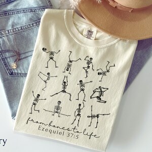 Op de afbeelding: Ivoor kleurig t-shirt met zwarte skelet illustraties in yoga poses. De tekst "from bones to life Ezekiel 37:5" is onderaan gedrukt. Het shirt wordt getoond met een blauwe jeans en een bruine hoed.