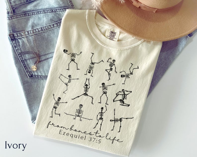 Op de afbeelding: Ivoor kleurig t-shirt met zwarte skelet illustraties in yoga poses. De tekst "from bones to life Ezekiel 37:5" is onderaan gedrukt. Het shirt wordt getoond met een blauwe jeans en een bruine hoed.