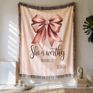 Puede incluir: Una manta a rayas rosas y blancas con una gran ilustración de lazo rosa. El texto "She is worthy" y "Proverbios 31" están escritos en una fuente cursiva. También se incluye el nombre "Olivia". La manta tiene un borde con flecos.