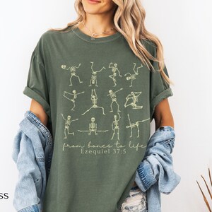 Op de afbeelding: Mosgroen t-shirt met skeletfiguren in yogahoudingen. De tekst "from bones to life" en "Ezequiel 37:5" is eronder gedrukt. Het shirt heeft een relaxte pasvorm en is gemaakt van zacht materiaal.