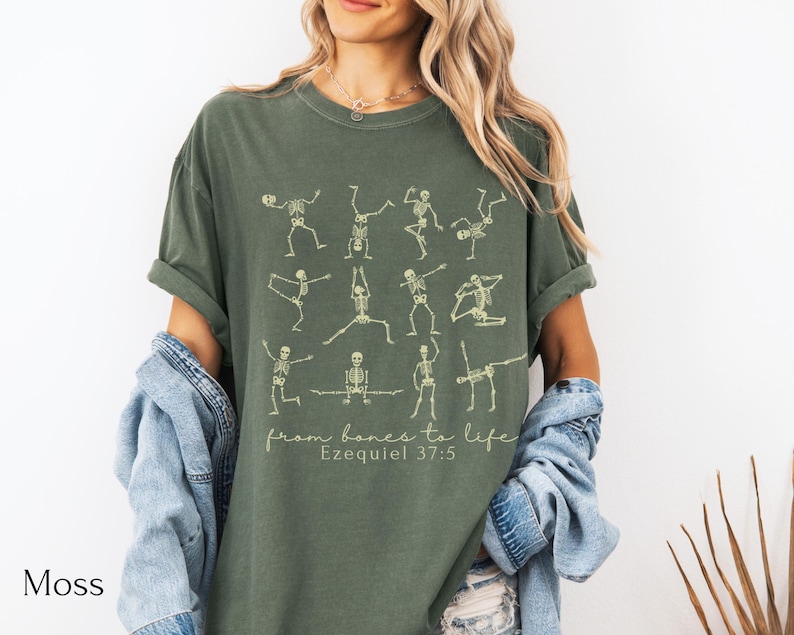Op de afbeelding: Mosgroen t-shirt met skeletfiguren in yogahoudingen. De tekst "from bones to life" en "Ezequiel 37:5" is eronder gedrukt. Het shirt heeft een relaxte pasvorm en is gemaakt van zacht materiaal.