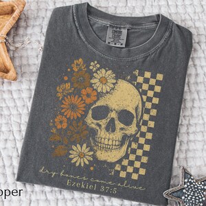 May include: Pepper-colored t-shirt featuring a skull design with floral accents in shades of orange and yellow. A checkered pattern is on the right side of the skull. The text "dry bones come alive Ezekiel 37:5" is printed below the skull.