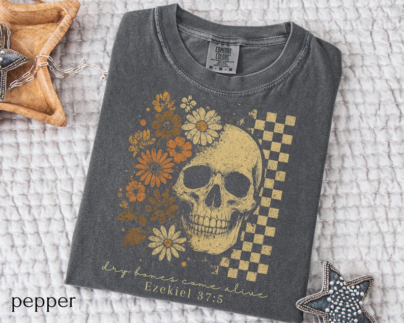 May include: Pepper-colored t-shirt featuring a skull design with floral accents in shades of orange and yellow. A checkered pattern is on the right side of the skull. The text "dry bones come alive Ezekiel 37:5" is printed below the skull.