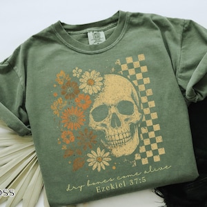 May include: Moss green t-shirt featuring a skull graphic with floral and checkered elements. The design includes the text "dry bones come alive" and "Ezekiel 37:5". The shirt is a comfortable, casual style.