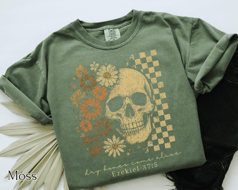 May include: Moss green t-shirt featuring a skull graphic with floral and checkered elements. The design includes the text "dry bones come alive" and "Ezekiel 37:5". The shirt is a comfortable, casual style.