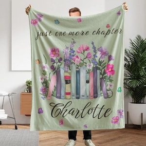 Puede incluir: Una manta de forro polar verde claro con una fila de libros coloridos coronados por flores rosas, moradas y blancas. El texto "just one more chapter" aparece en la parte superior y el nombre "Charlotte" en letra negra debajo.