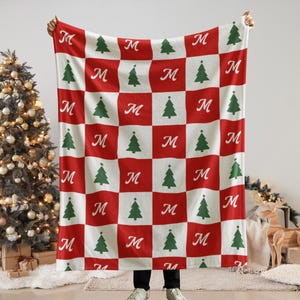 Puede incluir: Una manta rectangular con un patrón de cuadros rojos y blancos. Los cuadrados rojos contienen una letra M cursiva blanca, mientras que los cuadrados blancos presentan un gráfico de un árbol de Navidad verde. La manta se sostiene para mostrar el diseño.