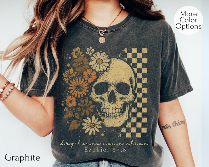 May include: Graphite-colored t-shirt featuring a skull design with floral accents and a checkered pattern. The design includes the text "dry bones come alive" and "Ezekiel 37:5". The shirt has a vintage, worn look.