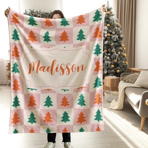 Puede incluir: Una manta rectangular con un patrón de cuadros repetitivo en rosa claro y blanco. La manta está decorada con iconos de árboles de Navidad que alternan entre verde y naranja y muestra el nombre Madisson en letra cursiva naranja.