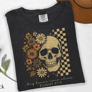 May include: Black t-shirt featuring a skull design with floral accents and a checkered pattern. The skull is surrounded by orange and cream-colored flowers. The text "dry bones come alive" and "Ezekiel 37:5" are printed on the shirt.