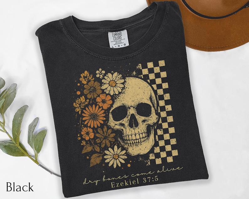 May include: Black t-shirt featuring a skull design with floral accents and a checkered pattern. The skull is surrounded by orange and cream-colored flowers. The text "dry bones come alive" and "Ezekiel 37:5" are printed on the shirt.
