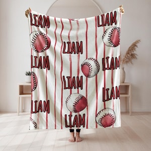 Puede incluir: Una manta blanca con un patrón de béisbol y rayas rojas. El nombre "LIAM" está impreso en rojo en la manta. La manta está sostenida por una persona.