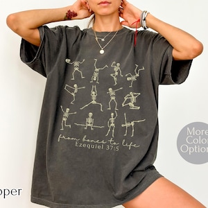Op de afbeelding: Pepperkleurig t-shirt met skeletfiguren in yogahoudingen. De tekst "from bones to life Ezekiel 37:5" staat onder de figuren. Het shirt heeft een relaxte pasvorm en een ronde hals.