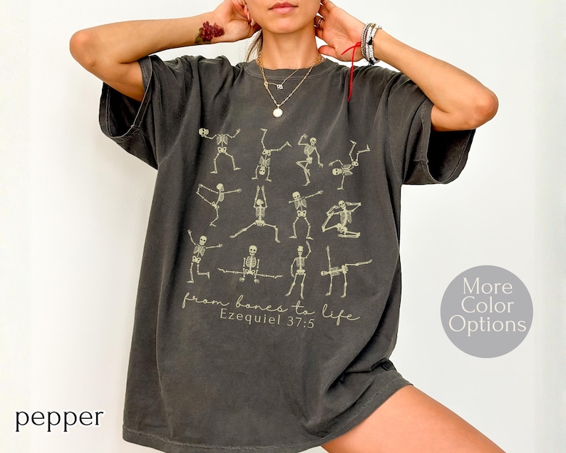 Op de afbeelding: Pepperkleurig t-shirt met skeletfiguren in yogahoudingen. De tekst "from bones to life Ezekiel 37:5" staat onder de figuren. Het shirt heeft een relaxte pasvorm en een ronde hals.