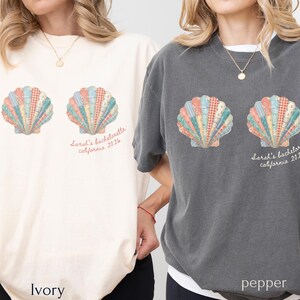 May include: Ivory and pepper-colored t-shirts featuring a seashell design with a patchwork pattern in pastel colors. The text "Sarah's bachelorette california 2026" is printed below the seashells. The shirts are casual and comfortable.