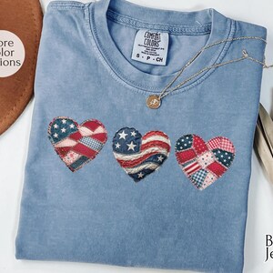 May include: A blue jean Comfort Colors t-shirt with three heart-shaped patches in red, white, and blue, featuring stars and stripes designs. A gold necklace with a pendant rests on the shirt. The shirt is size S-P-CH.