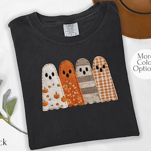 May include: Black t-shirt featuring four appliqued ghost figures in various patterns and colors. The ghosts have stitched details and are in white, orange, brown, and orange gingham. The shirt has the text "More Color Options" and "Black".