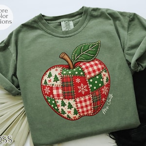 Puede incluir: Una camiseta de manga corta color verde musgo con un gráfico central de una manzana con diseño de patchwork de motivos navideños rojos y verdes, incluyendo árboles de Navidad, copos de nieve y cuadros. El texto Mrs. Smith está impreso.