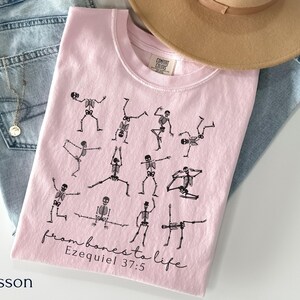 Op de afbeelding: Een lichtroze t-shirt met zwarte skeletfiguren in yogahoudingen. De tekst "from bones to life Ezekiel 37:5" is in zwarte letters gedrukt. Het shirt wordt getoond met een blauwe jeans en een beige hoed.