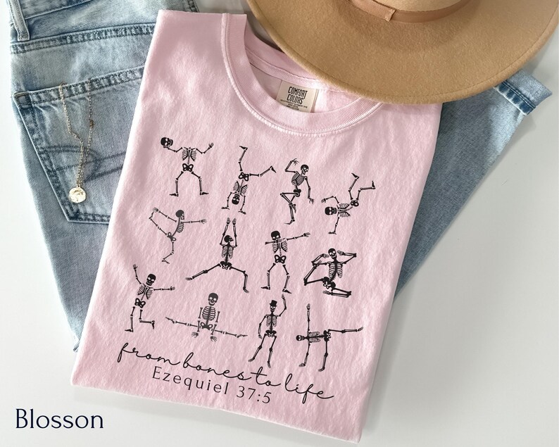 Op de afbeelding: Een lichtroze t-shirt met zwarte skeletfiguren in yogahoudingen. De tekst "from bones to life Ezekiel 37:5" is in zwarte letters gedrukt. Het shirt wordt getoond met een blauwe jeans en een beige hoed.