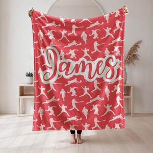 Puede incluir: Una manta roja con un patrón blanco de figuras atléticas en varias poses. El nombre "James" está escrito en una fuente grande y cursiva en el centro. La manta está sostenida por una persona.
