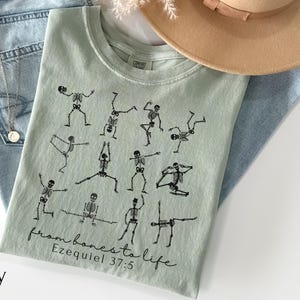 Op de afbeelding: Een lichtgroen t-shirt met zwarte skeletillustraties in verschillende yogahoudingen. De tekst "from bones to life" en "Ezequiel 37:5" zijn eronder gedrukt. Het shirt wordt gecombineerd met een blauwe jeans en een beige hoed.