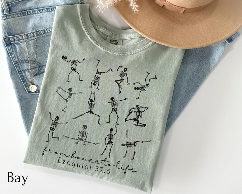 Op de afbeelding: Een lichtgroen t-shirt met zwarte skeletillustraties in verschillende yogahoudingen. De tekst "from bones to life" en "Ezequiel 37:5" zijn eronder gedrukt. Het shirt wordt gecombineerd met een blauwe jeans en een beige hoed.