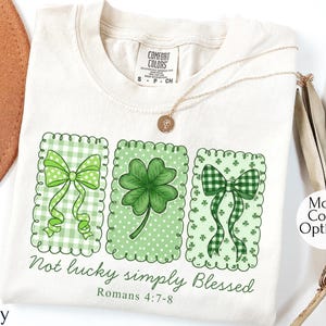 Puede incluir: Camiseta de color marfil con tres diseños verdes enmarcados, que incluyen lazos y un trébol de cuatro hojas. El texto "Not lucky simply Blessed" y "Romans 4:7-8" están impresos debajo. La camiseta tiene cuello redondo y mangas cortas.