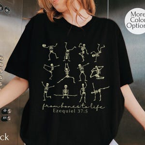 Op de afbeelding: Zwart T-shirt met een ontwerp van dansende skeletten in verschillende poses. De tekst "from bones to life" en "Ezequiel 37:5" zijn eronder gedrukt. Het shirt heeft een ronde hals en korte mouwen. De tekst "More Color Options" staat in een cirkel.