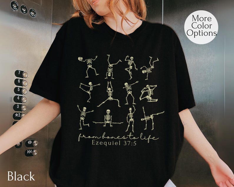 Op de afbeelding: Zwart T-shirt met een ontwerp van dansende skeletten in verschillende poses. De tekst "from bones to life" en "Ezequiel 37:5" zijn eronder gedrukt. Het shirt heeft een ronde hals en korte mouwen. De tekst "More Color Options" staat in een cirkel.