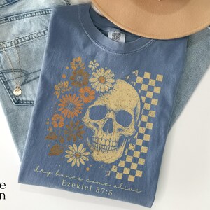 May include: A blue Comfort Colors t-shirt featuring a skull design with floral accents and a checkered pattern. The shirt includes the text "dry bones come alive" and "Ezekiel 37:5". The shirt is folded on top of blue jeans.