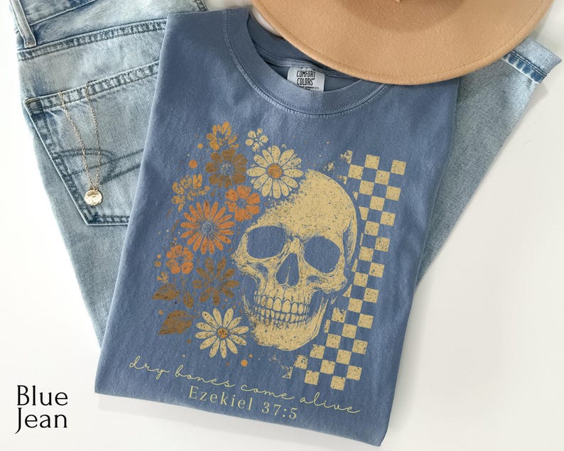 May include: A blue Comfort Colors t-shirt featuring a skull design with floral accents and a checkered pattern. The shirt includes the text "dry bones come alive" and "Ezekiel 37:5". The shirt is folded on top of blue jeans.