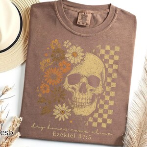 May include: Espresso-colored t-shirt featuring a skull design with floral accents and a checkered pattern. The graphic includes the text "dry bones come alive" and "Ezekiel 37:5". The shirt is a Comfort Colors XL-TG-BG.