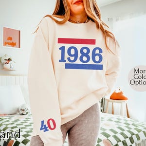 May include: Sand-colored sweatshirt with the year "1986" in blue, framed by red and blue bars. The number "40" is on the left sleeve in red and blue. The sweatshirt is a casual style.