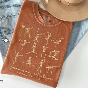 Op de afbeelding: Een oranje t-shirt met een ontwerp van yoga-houdingen met skeletten. De tekst "from bones to life" en "Ezequiel 37:5" is gedrukt. Het t-shirt wordt gecombineerd met een lichtblauwe jeans en een beige hoed.