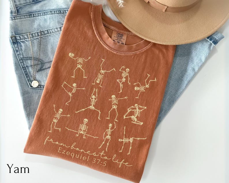 Op de afbeelding: Een oranje t-shirt met een ontwerp van yoga-houdingen met skeletten. De tekst "from bones to life" en "Ezequiel 37:5" is gedrukt. Het t-shirt wordt gecombineerd met een lichtblauwe jeans en een beige hoed.