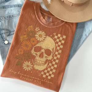 May include: Burnt orange t-shirt featuring a skull graphic with floral and checkered elements. The text "Dry bones come alive Ezekiel 37:5" is printed below the skull. The shirt is paired with light blue jeans and a tan hat.