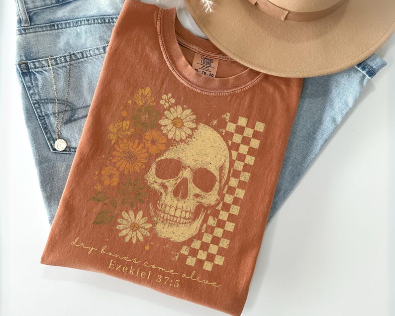 May include: Burnt orange t-shirt featuring a skull graphic with floral and checkered elements. The text "Dry bones come alive Ezekiel 37:5" is printed below the skull. The shirt is paired with light blue jeans and a tan hat.