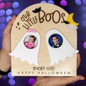 May include: A wooden photo frame shaped like a ghost with two photo cutouts. The frame is decorated with bats, stars, and a crescent moon. The text "Our Little Boos" is written on the frame. The text "Spooky Cute! Happy Halloween" is written below the photo cutouts.