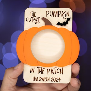 May include: A wooden pumpkin-shaped photo frame with a cutout for a picture. The frame is orange and has the text "The Cutest Pumpkin in the Patch Halloween 2024" on it.
