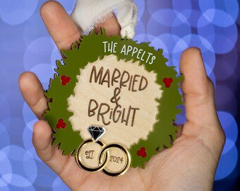 May include: A green wreath Christmas bauble with the text "The Appelts Married & Bright" and two gold rings with the dates "Est. 2023" and "2024" on them.