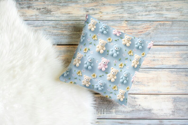 May include: A square pillow with a light blue background, covered in small teddy bear figures in pastel colors. The bears are beige, light blue, and pink, with small yellow flowers scattered around them. The pillow is on a wooden surface.