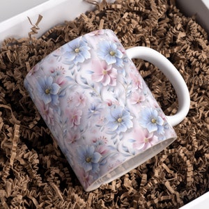 May include: A white ceramic mug with a floral design featuring pink and blue flowers on a white background. The mug is sitting in a brown paper shred box.