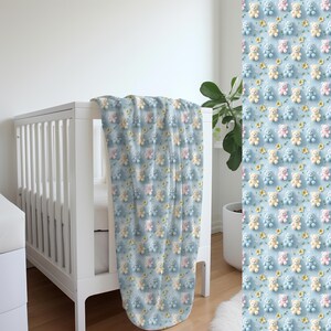 May include: A baby blanket with a repeating pattern of pastel-colored teddy bears and small yellow flowers on a light blue background. The blanket is draped over a white crib. The blanket is soft and cozy.
