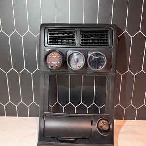 Puede incluir: Panel negro del salpicadero del coche con tres indicadores y rejillas de ventilación. Los indicadores incluyen un indicador de relación aire/combustible AEM. El panel tiene una abertura rectangular y un pequeño compartimento de almacenamiento con una perilla. El fondo es un patrón de baldosas geométricas en blanco y negro.