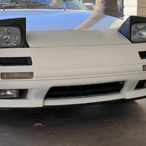 May include: A white Mazda RX-7 sports car with black headlights and a black grille. The car has a white body and black trim.