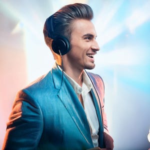 以下が含まれることがあります： 青いブレザーと白い襟付きシャツを着た男性が、右を見て笑顔を見せます。彼は耳に黒いヘッドホンをつけています。