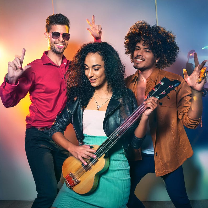以下が含まれることがあります： 男性2人と女性1人の3人が一緒に立っています。女性は茶色のアコースティックギターを持って演奏しています。左側の男性は赤いシャツを着ており、右側の男性は茶色のシャツを着ています。女性は黒い革のジャケットと緑色のスカートを着ています。