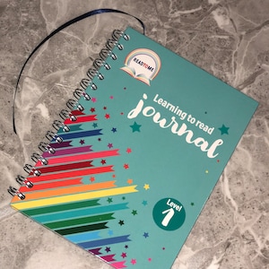 May include: A teal blue spiral bound journal with a rainbow design and stars. The cover has the text "Learning to read journal" and a logo that says "Read to Me". The journal is labeled "Level 1".