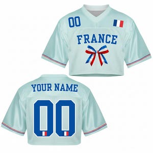 May include: Light blue cropped jersey with the word "FRANCE" in blue block letters, a blue, white, and red bow, and the French flag. The jersey has the number "00" on the front and back, and the words "YOUR NAME" on the back.